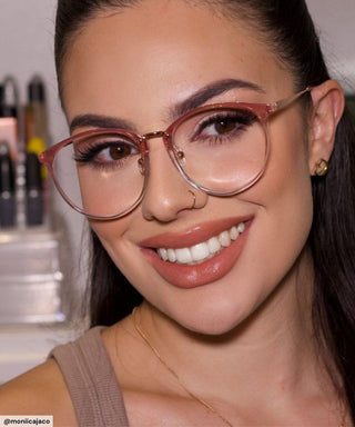 A woman with long dark hair smiles at the camera, wearing EyeCandys Infinity Oversized Oval pink eyeglasses, gold earrings, and a nose ring. Her makeup features defined brows, lashes, and nude lipstick. Makeup shelves are visible behind her.