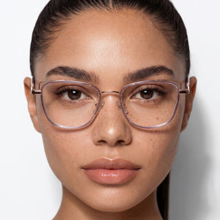 A woman with smooth skin and minimal makeup gazes at the camera, her brown eyes framed by EyeCandys Kaylee Cat Eye glasses. She has dark hair pulled back and stands against a plain white background, showcasing the stylish thin frames.
