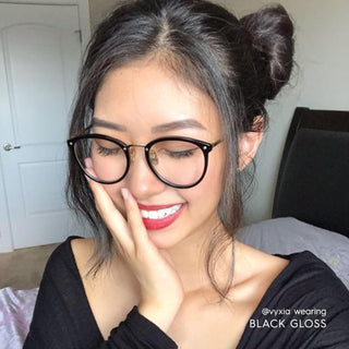 A young woman with dark hair in space buns smiles with her eyes closed, wearing EyeCandys Infinity Oversized Oval vintage eyeglasses in black. She touches her face with one hand. Text reads “@vyxia wearing BLACK GLOSS.”.