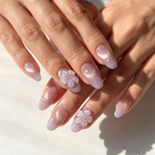 Hands with long, almond-shaped Lilac Dew Press-On Nails by EyeCandys in glossy pale pink. Two nails have 3D white floral designs and some feature raised pearl-like accents, giving a salon gel manicure appearance.