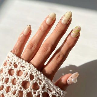 A hand with long, almond-shaped Little Bea Press-On Nails by EyeCandys features minimalist dots, delicate lines, and floral designs in neutral shades. A white crochet sleeve partially covers the hand.
