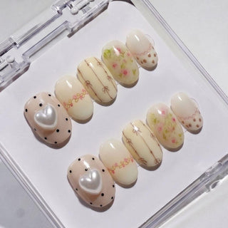 Little Bea Press-On Nails by EyeCandys feature pastel cottagecore designs—bows, tiny flowers, dots, scalloped edges, and two nails with large 3D pearl hearts—all handmade and arranged in a clear case for a salon gel manicure look.