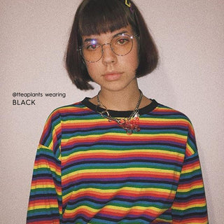 A person with short brown hair wears EyeCandys Luna Round eyeglass frames, a rainbow-striped shirt, and a chunky necklace, standing against an off-white wall. The text "@tteaplants wearing BLACK" appears on the left side.