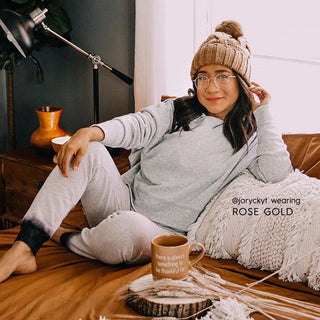 A woman in a cozy sweater, sweatpants, and knit beanie relaxes on rust-colored sheets, smiling as she adjusts her hat. She wears EyeCandys Luna Round titanium vintage-style glasses and holds a mug that reads, "There is always something to be thankful for.