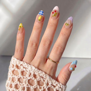 A hand in a crochet sweater displays EyeCandys’ Macedonia Press-On Nails—pastel, colorful designs like flowers, plaid, dots, a bow, and fruit art—each unique nail pops beside a simple gold ring.
