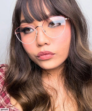 A young woman with long wavy brown hair and bangs models EyeCandys’ Madame Cat Eye lightweight metal frames, pairing them with a pink plaid top and neutral makeup as she gazes away from the camera with a calm expression.