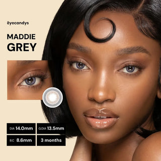 A woman with smooth dark skin and glossy nude lips wears Pink Label Maddie Grey contacts by EyeCandys. An inset shows her eye and a single lens, with text highlighting product details for these grey color contacts.