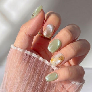 A hand with Matcha Made Press-On Nails by EyeCandys—manicured in pastel green, pearlescent shimmer, and translucent designs with tiny gold accents—partially covered by a sheer white fabric sleeve.