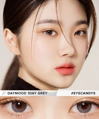 A close-up of a woman with fair skin, soft makeup, and straight dark hair showcases her grey contacts. Below her face is a zoom-in on the lens. Text: “Olola Daymood 1-Day Grey (10pk) #Olola.”.