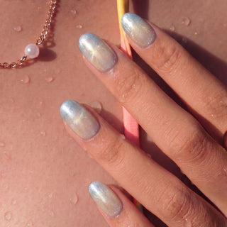 A hand with EyeCandys Moon Glass Press-On Nails in shiny, iridescent light blue rests on sun-kissed, wet skin. Water droplets and a delicate gold necklace with a pearl complete the look.