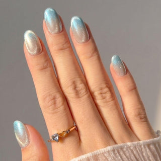 A hand features EyeCandys Moon Glass Press-On Nails in a shimmering blue and white gradient with an almond shape, complemented by a gold ring adorned with a heart-shaped gemstone.