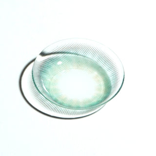 A single EyeCandys Pink Label Mallorca Blue contact lens, featuring a translucent blue with subtle green tint and dotted pattern, rests on a white surface and casts a soft shadow.
