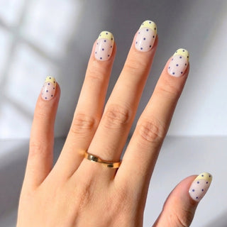 Close up hand showcasing Sunday Dots press on nails with soft off-white base, yellow French tips, and playful periwinkle dot design.