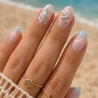 Wearing EyeCandys Pearl Jam Press-On Nails in ombre pastel blue and pink with iridescent flakes and a pearl nail featuring a starfish charm, the hand is set against sand and sea.