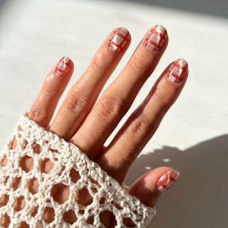 A hand with a white crocheted sleeve shows off EyeCandys Picnic Date Press-On Nails, featuring a red and white plaid design, glowing in natural sunlight.