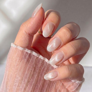 A hand with long, almond-shaped Seraphina Press-On Nails by EyeCandys features a sheer, shimmery white salon gel look. The wearer has a sheer light pink striped sleeve. Soft lighting highlights the nails' delicate elegance.