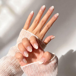 Close-up of hands wearing EyeCandys Seraphina Press-On Nails, featuring almond-shaped, milky white ombré tips. A sheer ribbed white long-sleeve top and soft sunlight highlight the elegant manicure.