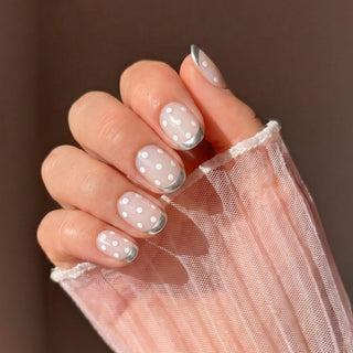 A hand with Silver Spotted Press-On Nails by EyeCandys—sheer pink salon gel nails featuring white polka dots and silver French tips—partly covered by sheer white mesh fabric.
