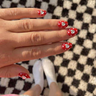 A hand wears EyeCandys' Spot the Difference Press-On Nails—red with white polka dots and tiny silver heart studs—posed against a black and white checkered background, with feet in white socks visible in the distance.