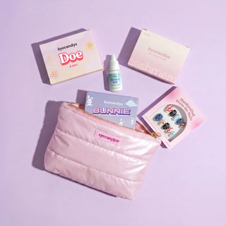 EyeCandys' The Complete Travel Pack, featuring a pink quilted pouch with the EyeCandys logo, displayed on a light purple surface alongside contact lens boxes, a bottle of contact lens solution, cat-themed lens cases, and a press-on nail set.