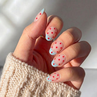 A hand wearing EyeCandys Unbothered Press-On Nails—light nude base, red polka dots, and light blue French tips—is shown in a cream knit sweater, inspired by a salon gel manicure.