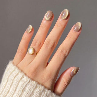 A hand wearing Velvet Taupe Press-On Nails by EyeCandys, shaped short and oval. The nails have holographic gold glitter. The person wears a gold ring with a white stone and a cream knit sweater against a plain gray background.