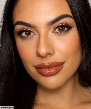 A woman with long dark hair and brown eyes wears EyeCandys Pink Label Vintage Grey lenses, looking at the camera. She has bold brows, long lashes, and warm natural makeup with glossy brown lipstick.