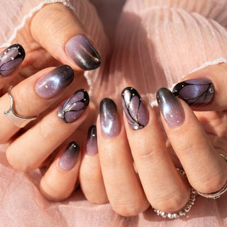Hands with long, almond-shaped Wing Woman Press-On Nails by EyeCandys featuring a shiny purple ombre with black tips, butterfly wing designs, white dots and gems. The person wears silver rings and a sheer pink fabric.