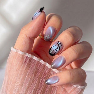 A hand with EyeCandys Wing Woman Press-On Nails in iridescent lavender, featuring black butterfly wing designs and gem accents on two nails, set against a soft background with a sheer pink sleeve.