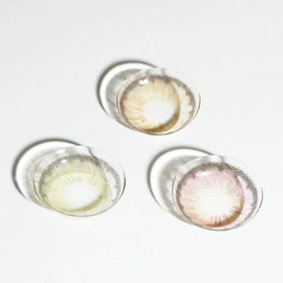 Three colored circle lenses, including EyeCandys Pink Label Angel Pink, plus brown and green patterned designs, are arranged on a white surface, each casting a soft shadow.