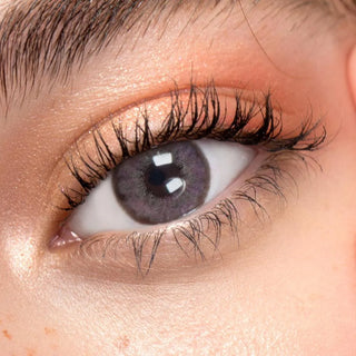 A close-up of an eye with long lashes, EyeCandys Pink Label Angel Pink contacts, and peach eyeshadow highlights detailed skin texture and expert makeup.