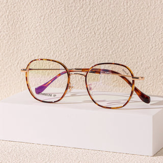 The EyeCandys Farrow Round glasses feature thin brown and gold rims with clear lenses in a vintage design. Resting on a white surface against a beige background, their narrow fit adds timeless elegance.