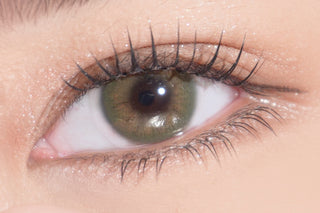 A close-up of a green hazel eye with long, defined eyelashes and shimmery makeup on the eyelid and surrounding skin. The skin appears light and smooth, and the eye is looking slightly to the right.