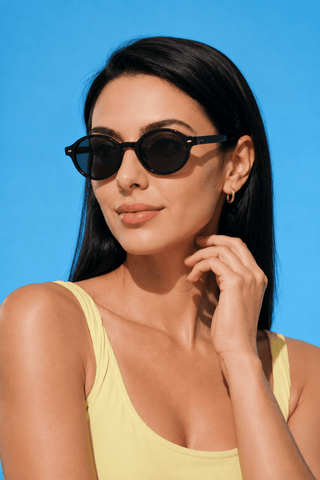 A woman with long dark hair, wearing EyeCandys Chase Round Oval glasses and a yellow sleeveless top, poses confidently against a bright blue background.