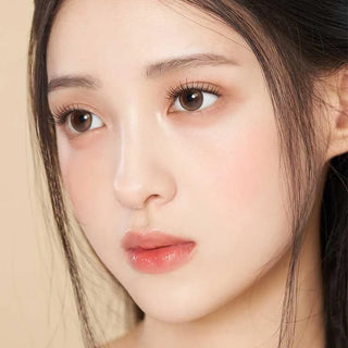 A close-up of a young woman with fair skin and straight dark hair wearing Gemhour Demeter 1-Day 13.5 Amber Brown contacts (10pk), which highlight her brown eyes, long lashes, and glossy pink lips as she gazes thoughtfully to the side.