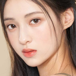 A close-up of a young woman with long dark hair, clear fair skin, and natural makeup wears Gemhour Demeter 1-Day 13.5 Amber Brown color contacts (10pk), gazing calmly at the camera against a neutral background.