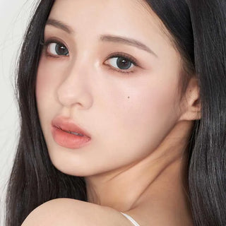 A young woman with long, straight dark hair and clear skin wears Gemhour Demeter 1-Day 13.0 Ocean Blue (10pk) color contacts by Gemhour. She has soft makeup, nude lips, a small beauty mark under her right eye, and a simple light background.