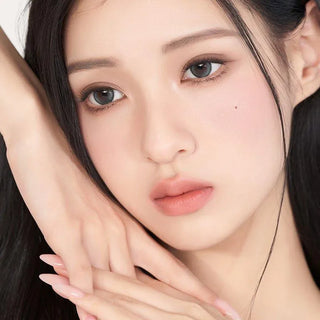 A woman with flawless, fair skin and long dark hair wears natural makeup and soft pink lips, serenely showcasing Gemhour Demeter 1-Day 13.0 Ocean Blue (10pk) color contacts by Gemhour, her hands gently resting near her face.