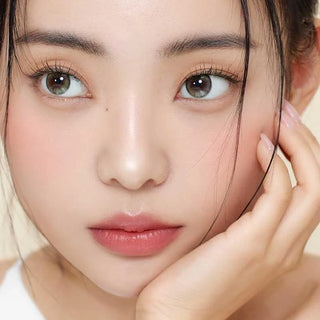 A young woman with smooth skin, natural makeup, and glossy lips gently touches her face. She wears Gemhour Demeter 1-Day 13.5 Olive Green contacts, which enhance her eyes as dark hair frames her face and she gazes to the side.