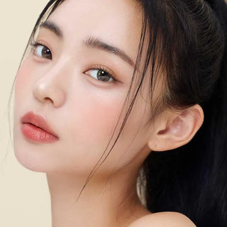 A young woman with smooth, clear skin and natural makeup wears Gemhour Demeter 1-Day 13.5 Olive Green color contacts, her straight black hair pulled back, soft pink lips, and defined brows set against a light beige background.