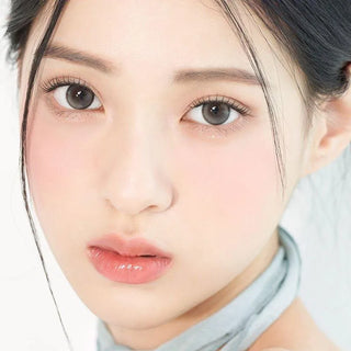 Close-up of a young woman with fair skin, soft makeup, and shiny grayish-brown eyes enhanced by Gemhour Demeter 1-Day 13.0 Pebble Grey (10pk) contact lenses. Her dark hair is pulled back and she wears a light blue top.