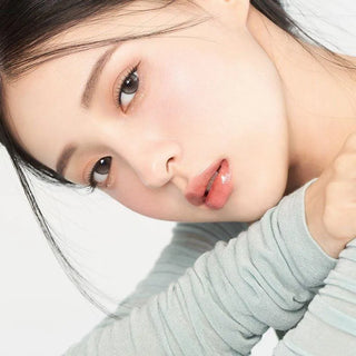 A young woman with clear skin and glossy lips gazes at the camera, resting her chin on her arms. She wears Gemhour Demeter 1-Day 13.0 Pebble Grey color contacts (10pk) by Gemhour, a light gray top, and has straight dark hair pulled back.