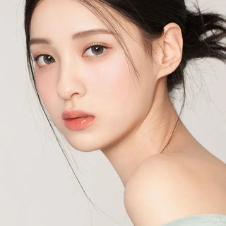 A young woman with fair skin and dark hair in a loose bun looks over her shoulder, wearing soft natural-toned makeup and glossy lips. Her calm expression is enhanced by Gemhour Demeter 1-Day 13.0 Pebble Grey contacts, set against a plain light background.