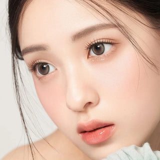 A young woman with smooth, fair skin and dewy makeup wears Gemhour Demeter 1-Day 13.0 Pebble Grey contact lenses, her light brown eyes beautifully enhanced as she gazes to the side, lips soft pink and hair pulled back.