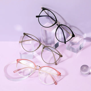 Three pairs of EyeCandys Infinity Oversized Oval eyeglasses—black, brown, and pink—are displayed on clear acrylic stands atop a pastel purple surface, with a glass sphere nearby, set against a soft lavender background.