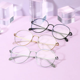 Three pairs of EyeCandys Luna Round eyeglass frames—in black, gold, and silver—are displayed on a pastel pink surface with transparent acrylic cubes, creating a subtle dark academia glasses vibe.