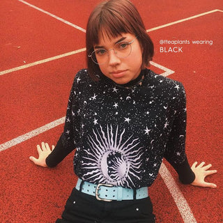 A person with short brown hair sits on a red track, wearing EyeCandys Luna Round glasses, a black shirt with white sun, moon, and star designs, black pants, and a light blue belt. Text reads "@tteaplants wearing BLACK.