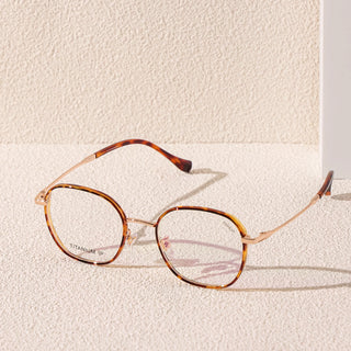 The EyeCandys Farrow Round glasses in tortoiseshell and gold with slim metal arms rest on a textured beige surface, their soft shadow cast in sunlight—a stylish, narrow-fit option for vintage round eyewear lovers.