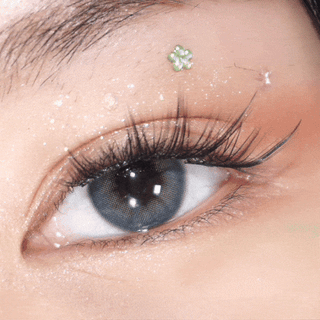 A close-up shot highlights long lashes and shimmery makeup, with a small green flower sticker on the eyelid. The eye, wearing EyeCandys Pink Label Mallorca Blue contacts, blinks slowly to enhance their natural-looking effect.