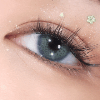 A close-up of a greenish-blue eye with long lashes, wearing EyeCandys Pink Label Amalfi Blue contacts. Glitter, stars, and dots on the eyelid enhance the lens’s soft, sparkling, and dreamy look.